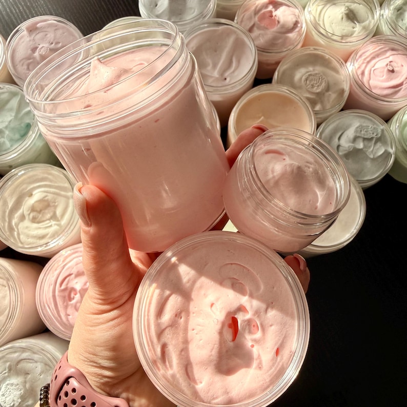 May include: A collection of clear plastic jars filled with thick, whipped cream-like substances in various pastel shades, including light pink, white, and pale green. The jars have smooth, rounded edges and are filled to the top with textured product.