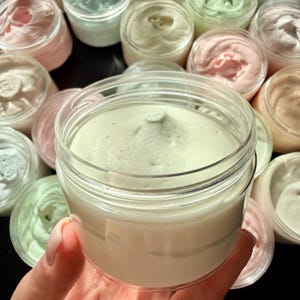 May include: A clear plastic jar filled with pale mint green whipped cream, featuring a smooth, swirled texture with a peak at the center. The container is transparent, revealing the thick, airy consistency of the product inside.