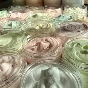 May include: A collection of clear plastic jars filled with whipped body butter in various pastel shades including light pink, mint green, pale yellow, and soft lavender. Each jar features a swirled, textured surface of the cream.