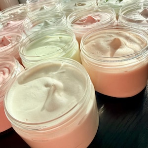 May include: A collection of clear plastic jars filled with whipped body butter in various pastel shades including soft pink, pale peach, and light mint green. Each jar features a smooth, swirled top surface, showcasing the creamy texture.