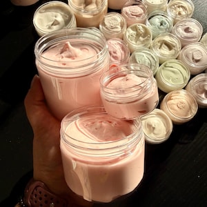 May include: A collection of clear plastic jars filled with thick, whipped cream-like substances in various pastel shades including light pink, soft green, and off-white. The jars are arranged in a group, with some held in the foreground.