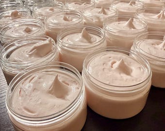 May include: A collection of small, clear jars filled with a light pink, whipped cream-like substance. Each jar has a silver lid and a dollop of the pink cream is piped on top. The jars are arranged in rows, suggesting a skincare product.