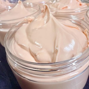 May include: A clear plastic jar filled with a thick, smooth, peach-colored cream. The cream is swirled into a peak at the center, extending slightly above the rim of the container. The texture appears dense and creamy, with a soft, matte finish.