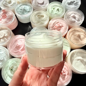May include: A collection of round, clear plastic jars filled with thick, whipped body butter in various pastel shades including white, pale pink, mint green, and soft peach. Each jar features a smooth, swirled top surface of the cream.