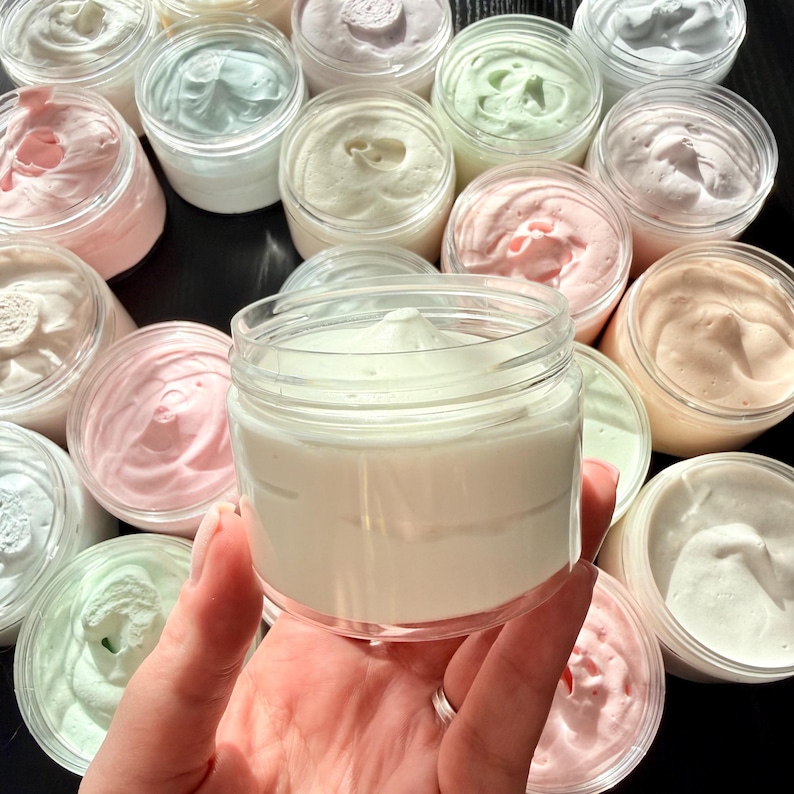 May include: A collection of round, clear plastic jars filled with thick, whipped body butter in various pastel shades including white, pale pink, mint green, and soft peach. Each jar features a smooth, swirled top surface of the cream.