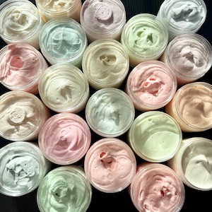 May include: A collection of round, clear plastic jars filled with whipped body butter in various pastel shades including soft pink, mint green, pale yellow, and white. Each jar features a textured, swirled surface on top of the cream.