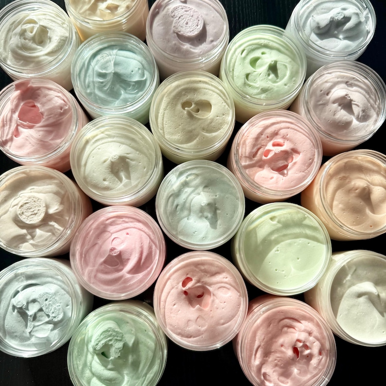 May include: A collection of round, clear plastic jars filled with whipped body butter in various pastel shades including soft pink, mint green, pale yellow, and white. Each jar features a textured, swirled surface on top of the cream.