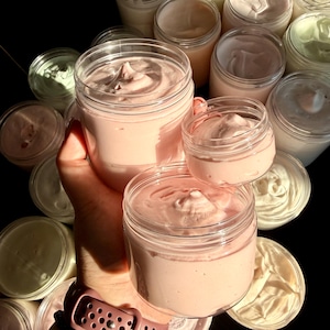 May include: A collection of clear plastic jars filled with thick, whipped cream-like substances in shades of pale pink, white, and light green. The jars have smooth, rounded tops and are arranged in a cluster, showcasing the textured, swirled surfaces.
