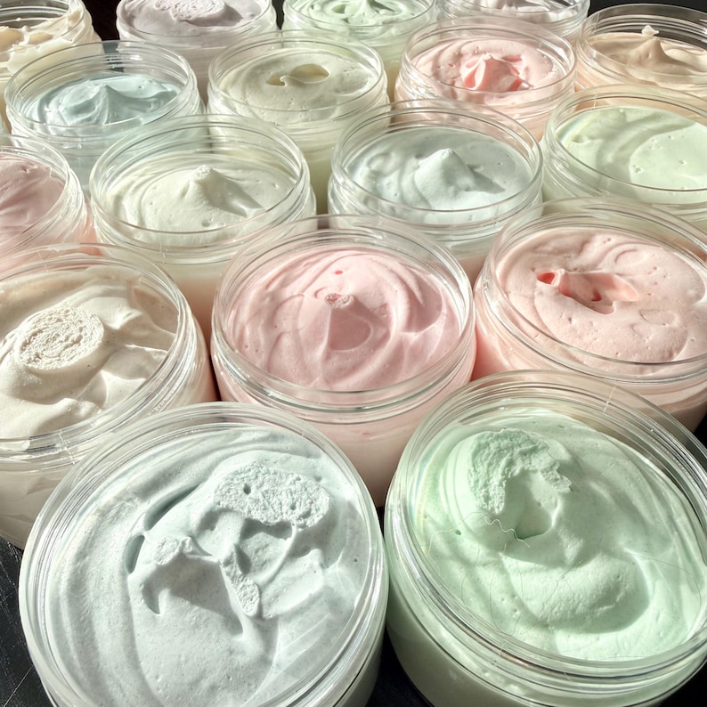 May include: A collection of clear plastic jars filled with whipped body butter in various pastel shades including light pink, mint green, pale blue, and off-white. Each jar features a thick, creamy texture with visible swirls and peaks on top.