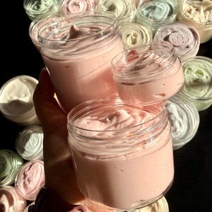 May include: A collection of clear plastic jars filled with whipped body butter in various pastel colors including light pink, mint green, and cream. The jars feature smooth, swirled textures on the surface of the cream, with several jars arranged in a cluster.