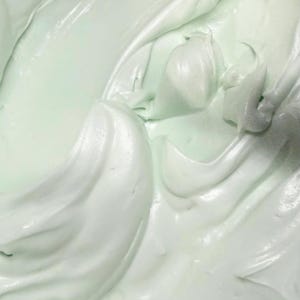 May include: A thick, creamy substance with a smooth, glossy texture and a pale mint green color. The surface features soft, swirling peaks and deep folds, creating a dense, whipped appearance with subtle highlights reflecting light across the curves.