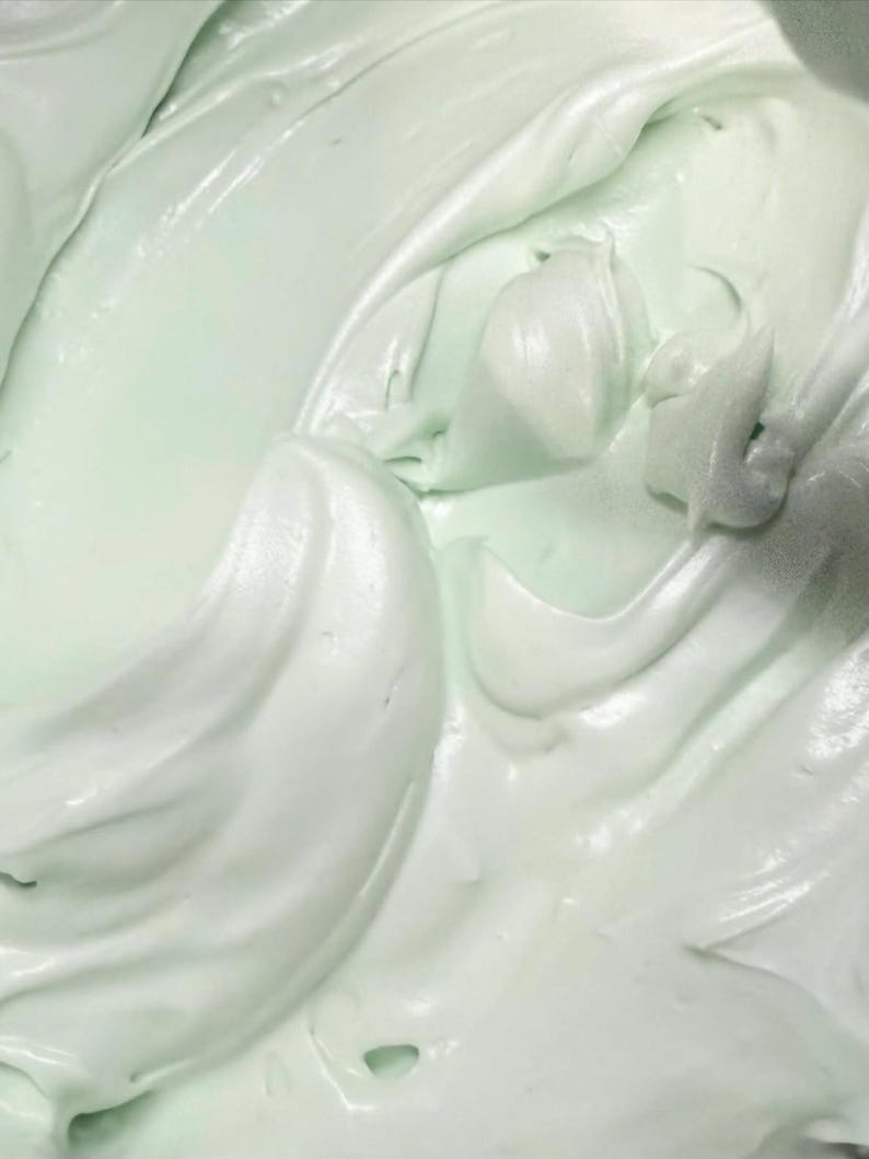 May include: A thick, creamy substance with a smooth, glossy texture and a pale mint green color. The surface features soft, swirling peaks and deep folds, creating a dense, whipped appearance with subtle highlights reflecting light across the curves.