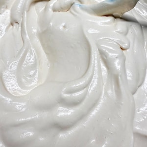 May include: A thick, creamy, off-white substance with a smooth, glossy texture and swirling patterns. The mixture appears dense and aerated, showing soft peaks and gentle folds throughout the surface, consistent with a whipped batter or frosting.