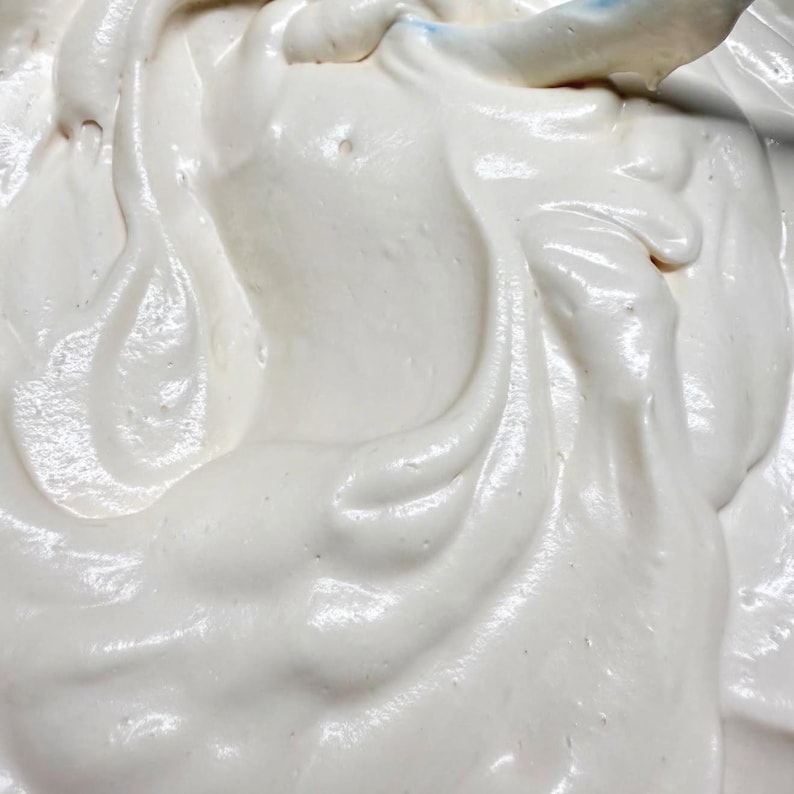 May include: A thick, creamy, off-white substance with a smooth, glossy texture and swirling patterns. The mixture appears dense and aerated, showing soft peaks and gentle folds throughout the surface, consistent with a whipped batter or frosting.
