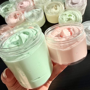 May include: A collection of clear plastic jars filled with whipped cream in various pastel colors including mint green, soft pink, pale yellow, and white. Each jar features a smooth, swirled top texture, showcasing the thick consistency of the product.