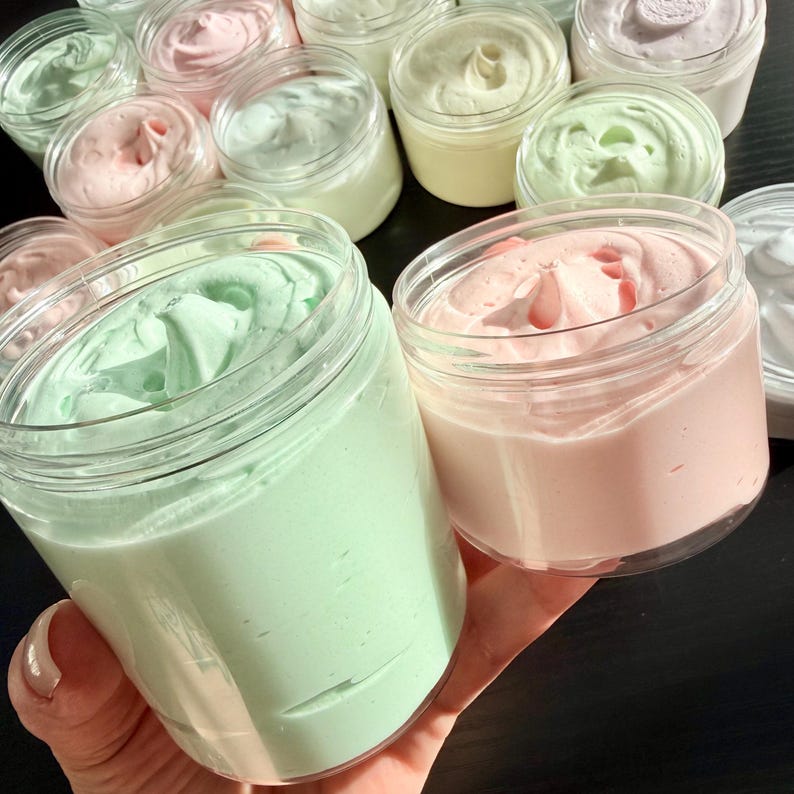 May include: A collection of clear plastic jars filled with whipped cream in various pastel colors including mint green, soft pink, pale yellow, and white. Each jar features a smooth, swirled top texture, showcasing the thick consistency of the product.
