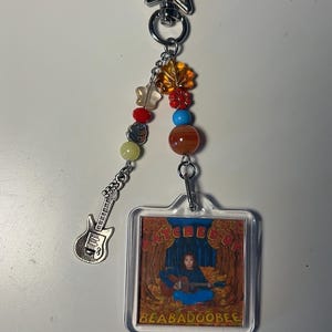 May include: A keychain with a star-shaped clasp and a clear, rectangular charm featuring a person playing a guitar. The charm is adorned with colorful beads and a silver guitar charm. The text "BEABADOOBEE" is visible.