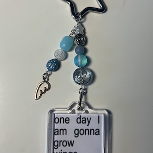 May include: A keychain featuring a silver star, blue beads, and a clear rectangular tag. The tag reads "one day I am gonna grow wings". A gold-coloured wing charm is also attached.