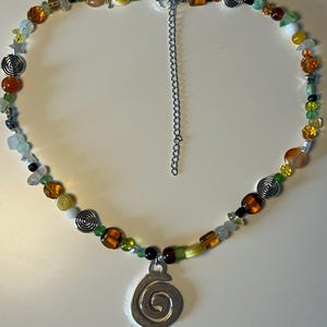 May include: A colorful beaded necklace featuring a variety of beads in shades of orange, green, yellow, and clear. The necklace includes silver spiral charms and a larger spiral pendant. The necklace is secured with a silver chain.