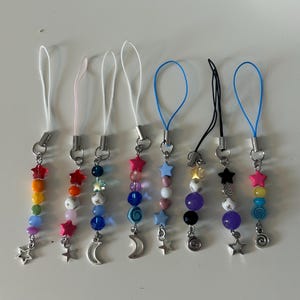 May include: A selection of beaded phone charms. Each charm has a loop, metal clasp, and colourful beads. The charms feature star, moon, and spiral designs. The beads are in shades of pink, blue, yellow, and purple. The charms are for mobile phones.