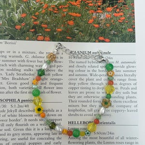 Puede incluir: Una pulsera de cuentas coloridas con cuentas verdes, amarillas, naranjas y blancas, incluyendo elementos en forma de flor. La pulsera se muestra sobre una página impresa.