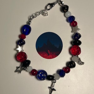 Puede incluir: Un brazalete de plata con cuentas rojas, azules, blancas y negras. Las cuentas tienen estrellas y lunas. El brazalete tiene un cierre de plata.