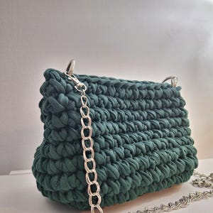 May include: A dark green, handmade crochet bag with a silver chain strap. The bag has a rectangular shape with rounded edges and a textured, woven appearance. The silver chain is attached to the bag with silver clasps.