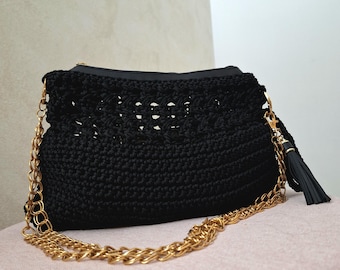 Könnte beinhalten: Schwarze Häkeltasche mit goldener Kette und schwarzem Quastenanhänger. Die Tasche hat einen Reißverschluss und ein offenes Webmuster. Die goldene Kette ist über die Vorderseite der Tasche drapiert.