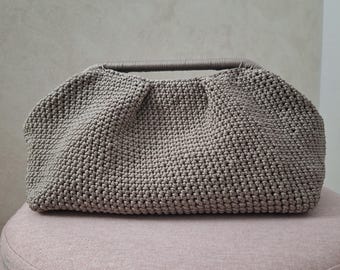 Könnte beinhalten: Eine handgehäkelte Handtasche in Taupe mit runder Form und passendem Griff. Die Tasche liegt auf einem rosa, abgerundeten Hocker. Der Häkelstich erzeugt eine strukturierte Oberfläche.