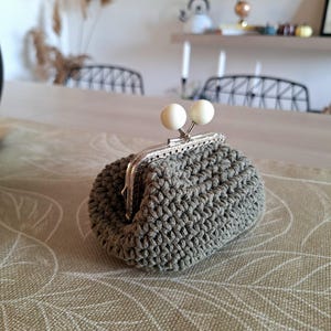 Peut inclure: Un petit porte-monnaie crocheté gris avec un fermoir argenté et deux accents sphériques blancs. Le sac est fait d'un matériau tissé texturé et repose sur une surface de ton neutre. Le fermoir est ouvert, révélant l'intérieur.