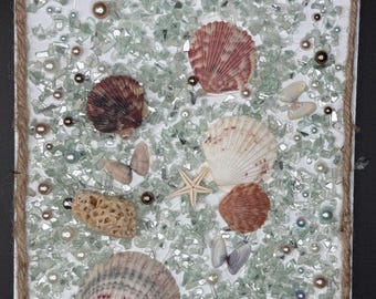 Può includere: Un'opera d'arte murale rettangolare con sfondo bianco ornato da conchiglie, pezzi di vetro e perle. Le conchiglie sono di vari colori e forme, con una stella marina e piccoli accenti di farfalle. L'opera è incorniciata con corda naturale.