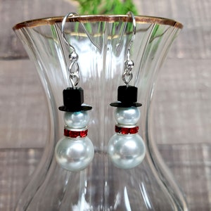 May include: A pair of silver snowman earrings with white pearl beads, black hats, and red rhinestone accents.