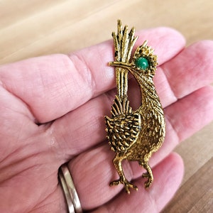 Peut inclure: Une broche dorée en forme de coureur des routes avec un œil en pierre précieuse verte. L'oiseau est debout sur une patte, les ailes déployées.