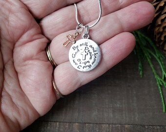 May include: Silver necklace with a round pendant that reads "Be still and know that I am God - Psalm 46:10" and a small gold cross charm.