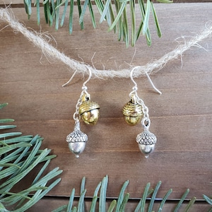 May include: A pair of silver dangle earrings with gold acorn charms. The earrings are hanging from a piece of twine against a wood background.