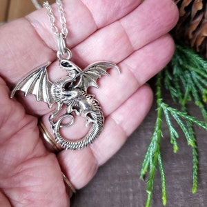 May include: Silver dragon pendant necklace. The dragon features detailed wings, scales, and a curled tail. The pendant hangs from a delicate silver chain. The necklace is held in a hand, with a natural background.