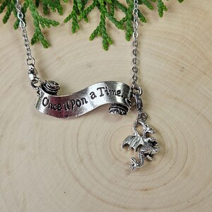 May include: A silver necklace with a banner inscribed with "Once Upon a Time" and a dragon pendant. The banner has rose detailing. The necklace is suspended from a silver chain, with green foliage in the background.