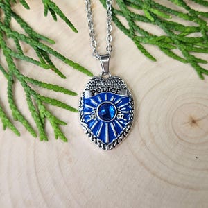 May include: A silver-toned police badge pendant necklace. The blue enamel badge features the word "POLICE" and a central blue gemstone. The necklace is displayed on a wooden surface with green foliage.