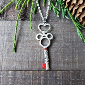 A silver necklace, the pendant is an antique silver key hanging from a silver heart.  The key has mouse ears, and a red M at the bottom.