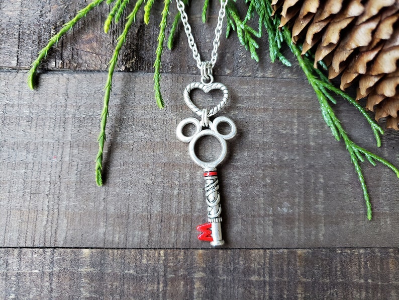 A silver necklace, the pendant is an antique silver key hanging from a silver heart.  The key has mouse ears, and a red M at the bottom.