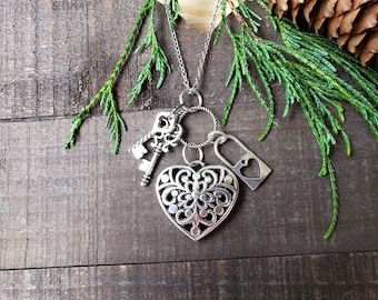 May include: Silver necklace with a heart-shaped pendant, a key charm, and a padlock charm. The necklace is on a wooden background with green foliage.