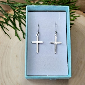 May include: A pair of silver cross earrings displayed in a light blue box. The earrings have a simple, elegant design with a cross shape. The box is open, revealing the earrings against a gray background. The background includes wood and greenery.