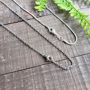 K&ouml;nnte beinhalten: Zwei silberne Halsketten mit unterschiedlichen Kettenstilen, eine zarte Schlangenkette und die andere eine robustere Panzerkette. Beide haben Karabinerverschl&uuml;sse. Die Halsketten werden auf einer rustikalen Holzoberfl&auml;che mit gr&uuml;nen Farnakzenten pr&auml;sentiert.