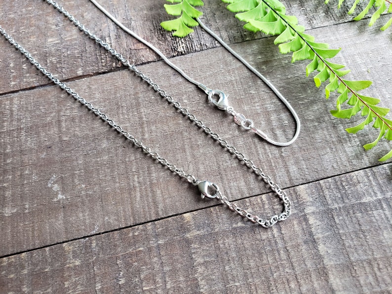 K&ouml;nnte beinhalten: Zwei silberne Halsketten mit unterschiedlichen Kettenstilen, eine zarte Schlangenkette und die andere eine robustere Panzerkette. Beide haben Karabinerverschl&uuml;sse. Die Halsketten werden auf einer rustikalen Holzoberfl&auml;che mit gr&uuml;nen Farnakzenten pr&auml;sentiert.