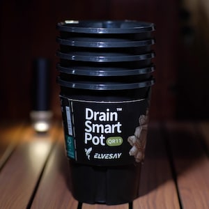 May include: A stack of black plastic plant pots with the text "Drain Smart Pot" and "QR11" on the label. The pots are sitting on a wooden surface. The pots are designed for drainage.