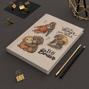 May include: A beige journal with illustrations of cartoon monkeys hugging. The journal cover has the text "You are stronger than you think" and "Be Brave" in a handwritten style. Gold and black pencils and binder clips are also visible.