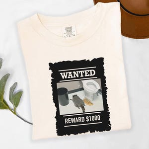 May include: Off-white t-shirt with a black and white graphic depicting a raccoon in a bathroom, with the words "WANTED" and "REWARD £1000". The shirt is folded, and a brown hat is visible in the background. The t-shirt is displayed on a white surface.