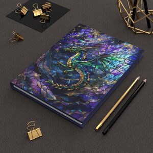 May include: A hardbound journal with a stained-glass style dragon design. The cover features a green and blue dragon against a backdrop of purple, blue, and gold. The journal is rectangular with a dark blue border.