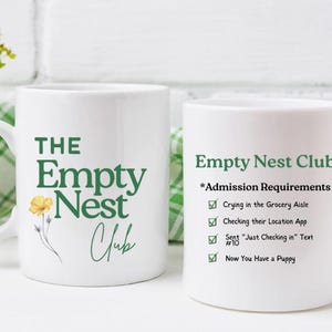 Puede incluir: Dos tazas de cerámica blancas. Una taza tiene "The Empty Nest Club" en texto verde con una ilustración de flor amarilla. La otra taza tiene "Empty Nest Club" y requisitos de admisión en texto verde.