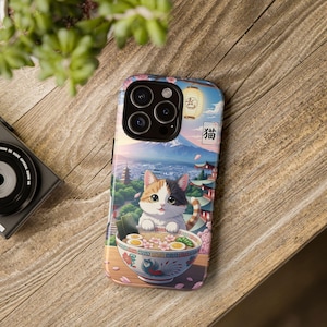 Puede incluir: Funda de teléfono con una colorida ilustración de un gato calicó en un cuenco de ramen. El fondo representa un paisaje japonés con el monte Fuji, pagodas y flores de cerezo. El carácter japonés para "gato" es visible.